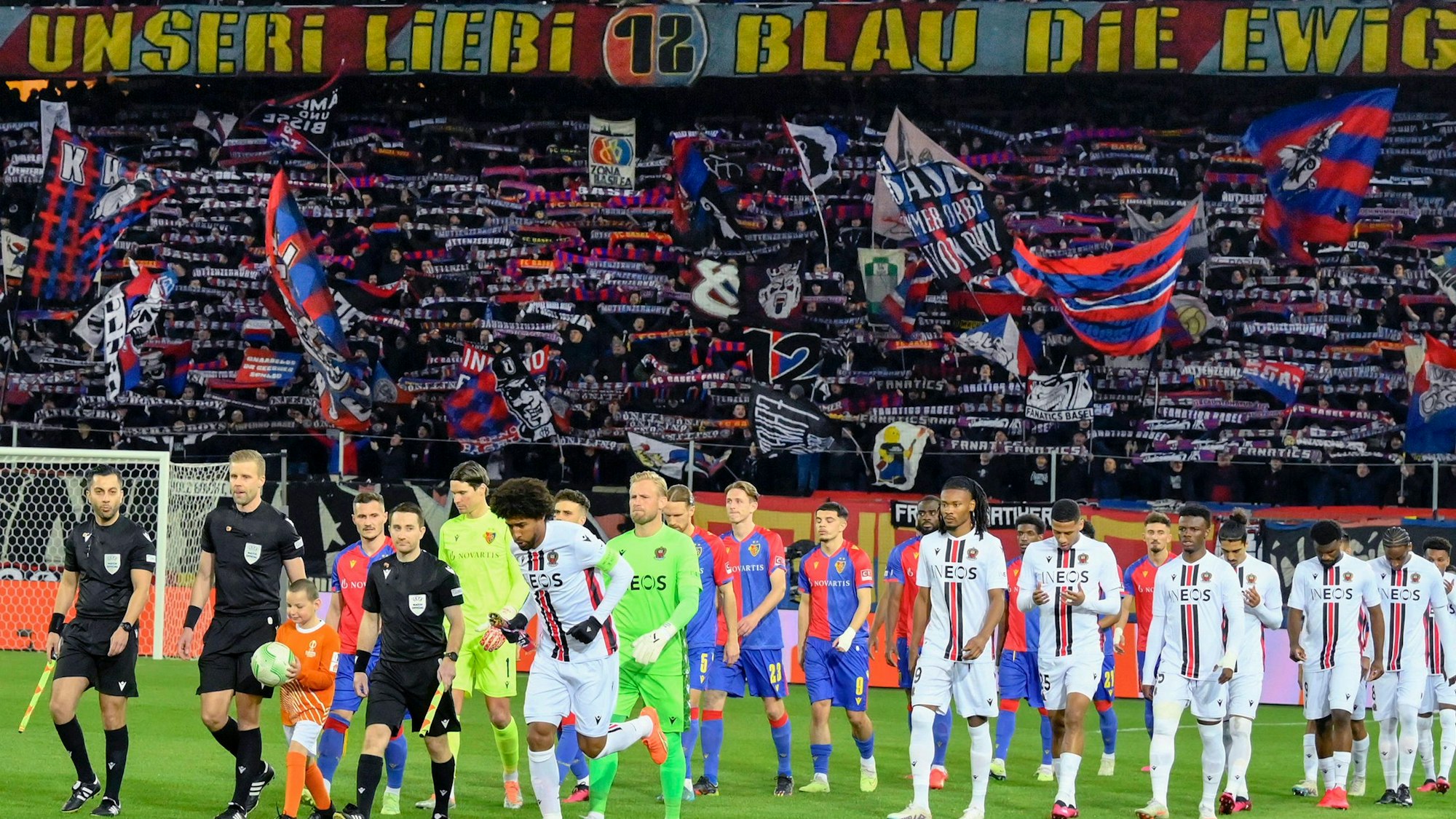Angeführt vom Schiedsrichtergespann, laufen zwei Fußball-Mannschaften ins Stadion ein. Im Hintergrund sieht man die Fan-Tribüne, wo Fans ihre Schals und Fahnen schwenken.