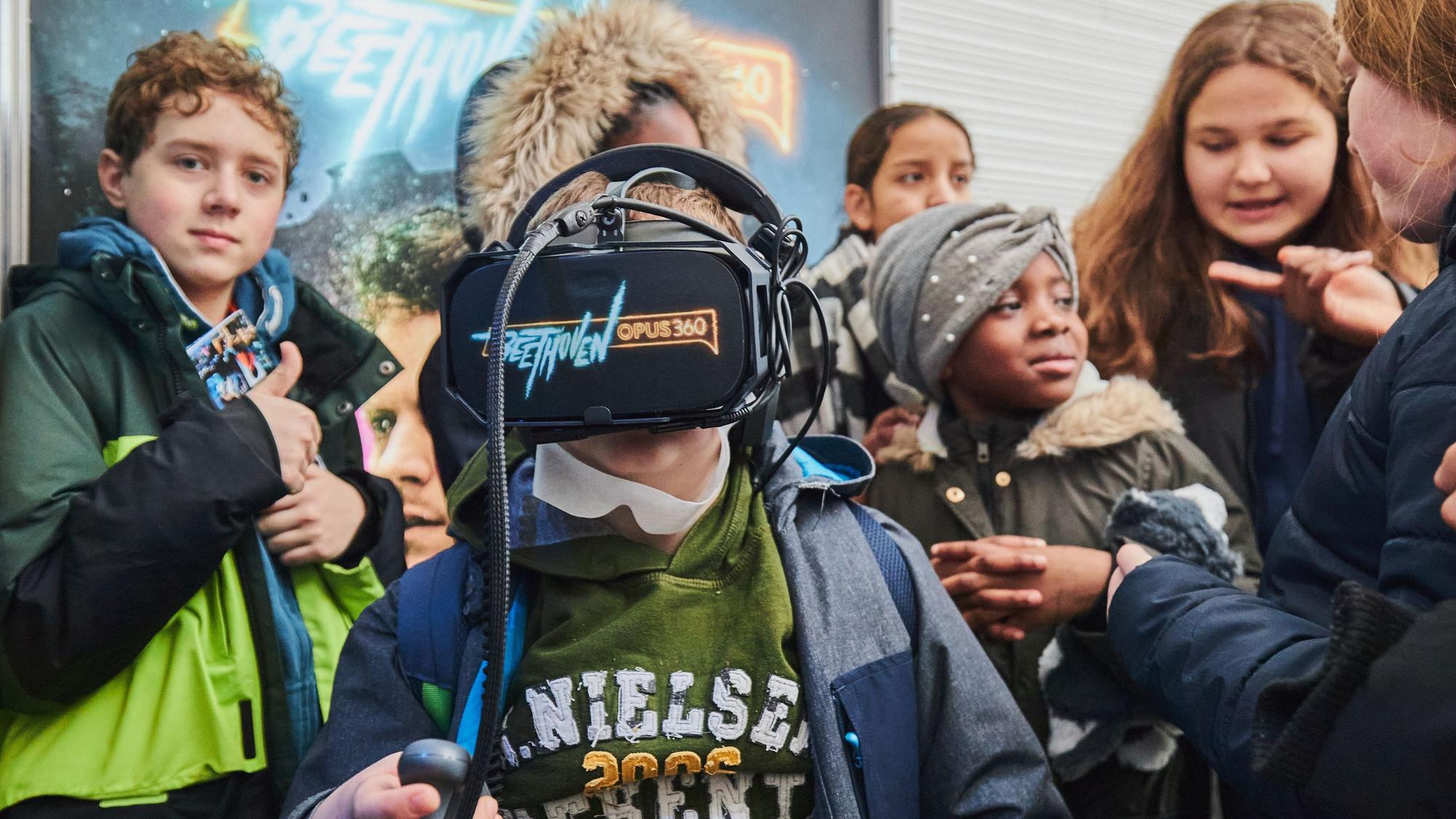 Ein Junge hat eine VR-Brille auf, auf der „Beethoven Opus 360 “steht. Um ihn herum haben sich Mitschülerinnen und Mitschüler versammelt.