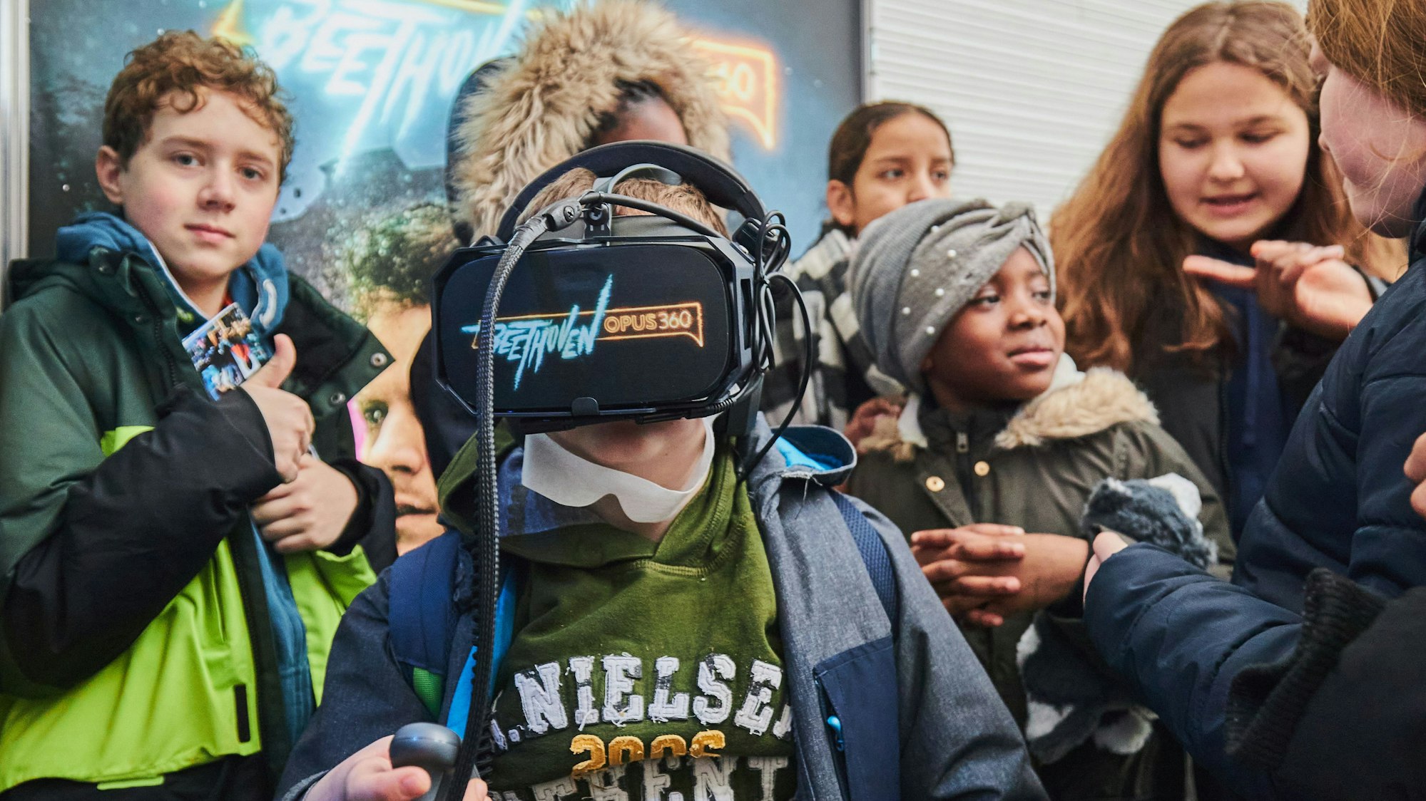 Ein Junge hat eine VR-Brille auf, auf der „Beethoven Opus 360 “steht. Um ihn herum haben sich Mitschülerinnen und Mitschüler versammelt.