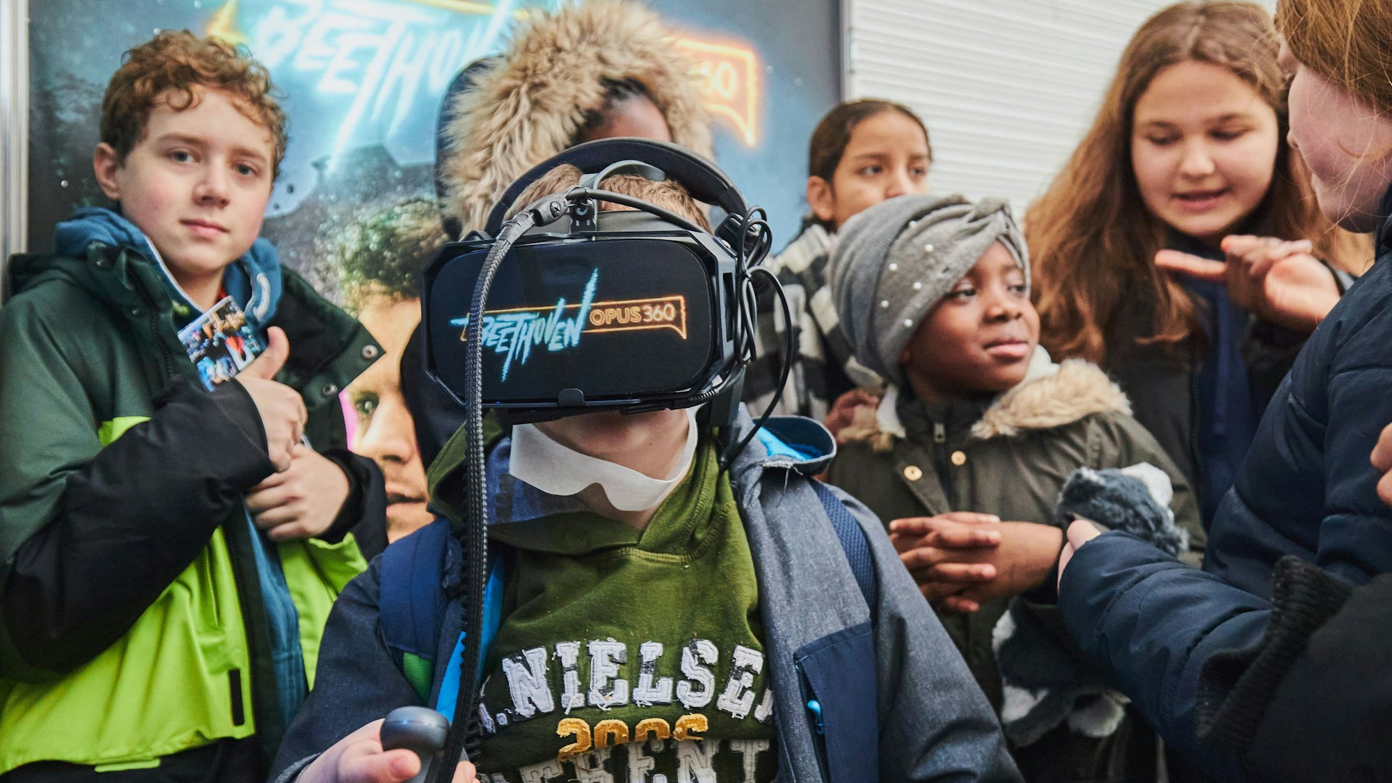 Ein Junge hat eine VR-Brille auf, auf der „Beethoven Opus 360 “steht. Um ihn herum haben sich Mitschülerinnen und Mitschüler versammelt.