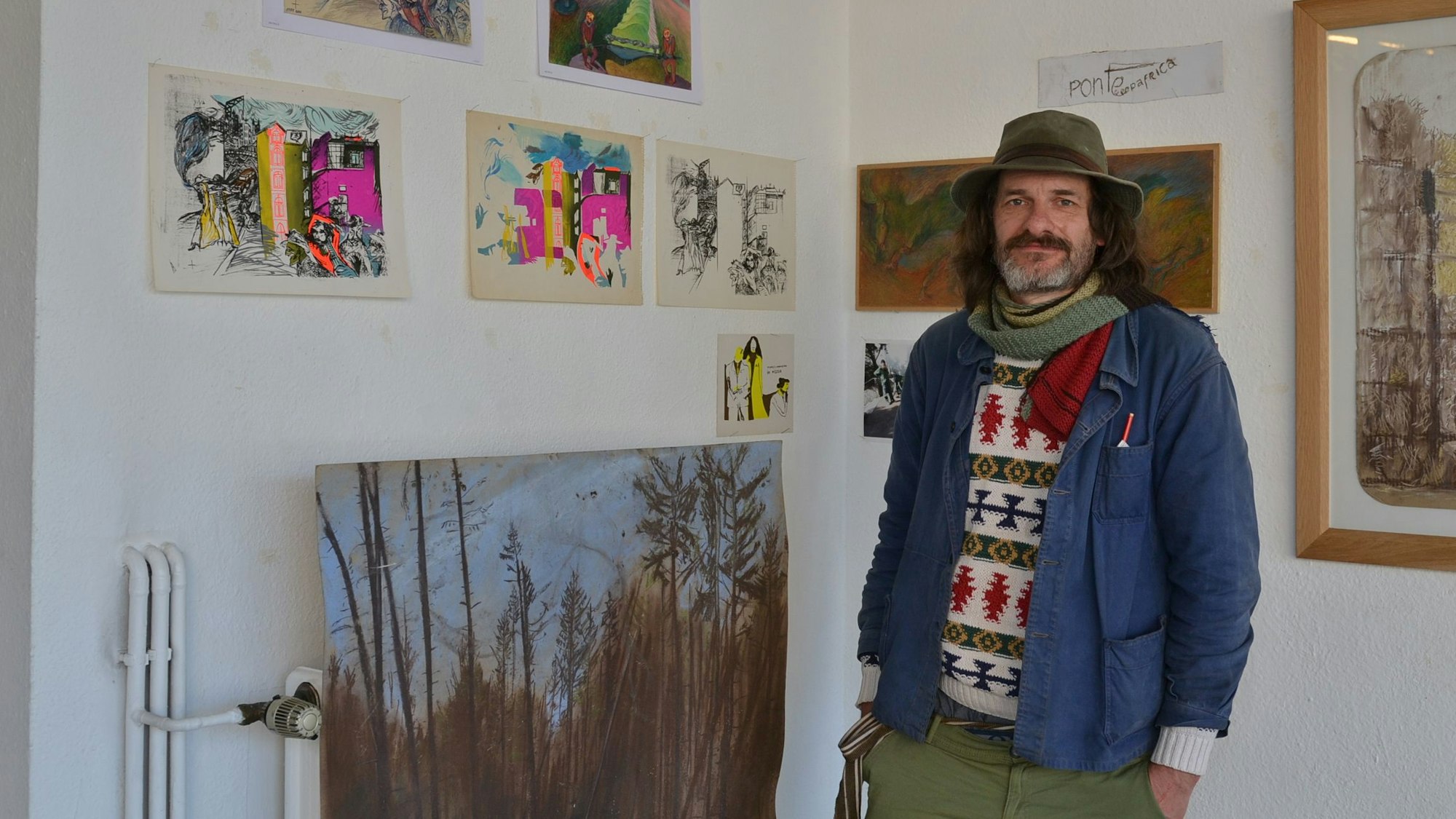 Der Künstler steht vor seinen Werken. Er trägt einen bunten Strickpulli, eine blaue Jacke und einen Hut.