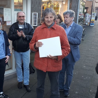 Ein junger Mann steht neben einer älteren Frau, die einen Ordner in der Hand hält. Daneben sind weitere Personen zu sehen. Alle stehen auf einem Bürgersteig vor einer Apotheke.