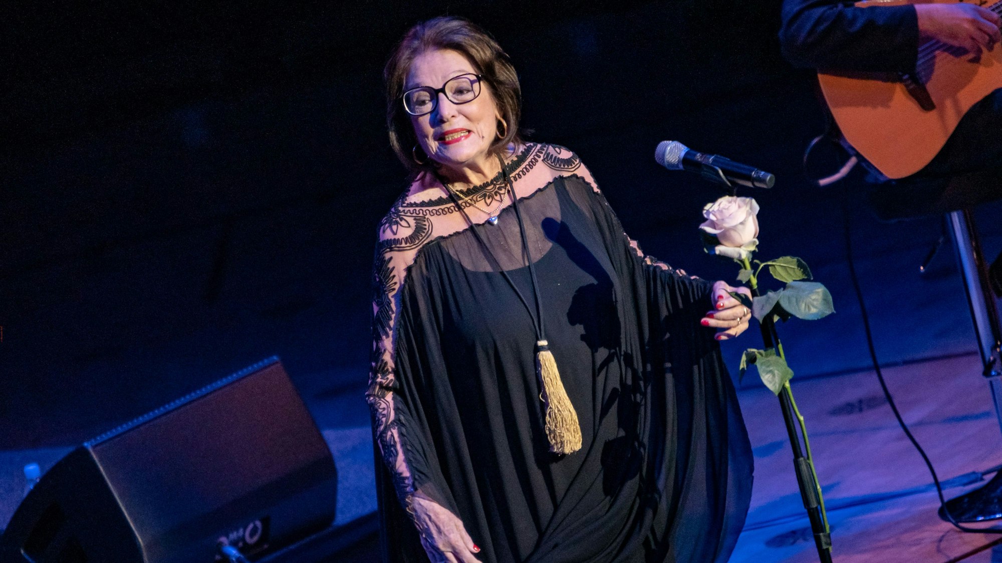 Nana Mouskouri im Oktober 2018 bei ihrem letzten Kölner Konzert in der Philharmonie. Sie steht vor dem Mikro mit einer Rose. Sie trägt ein langes schwarzes Kleid.