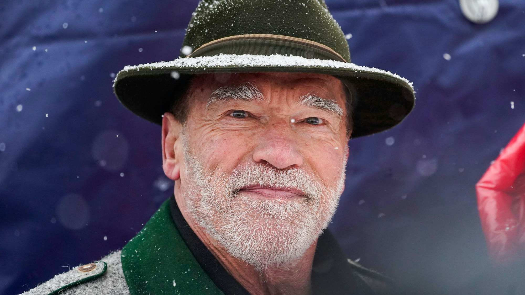 Arnold Schwarzenegger mit Trachtenhut. Es schneit um ihn herum.