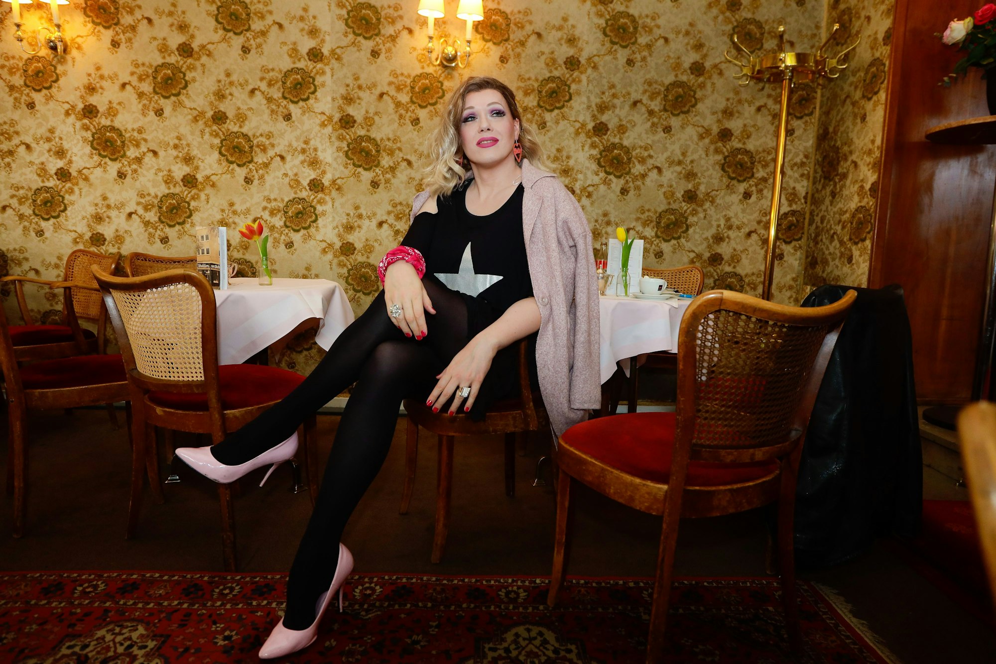 Cassy Carrington sitzt mit überschlagenen Beinen im Café Wahlen. Sie guckt süffisant oben an der Kamera vorbei.