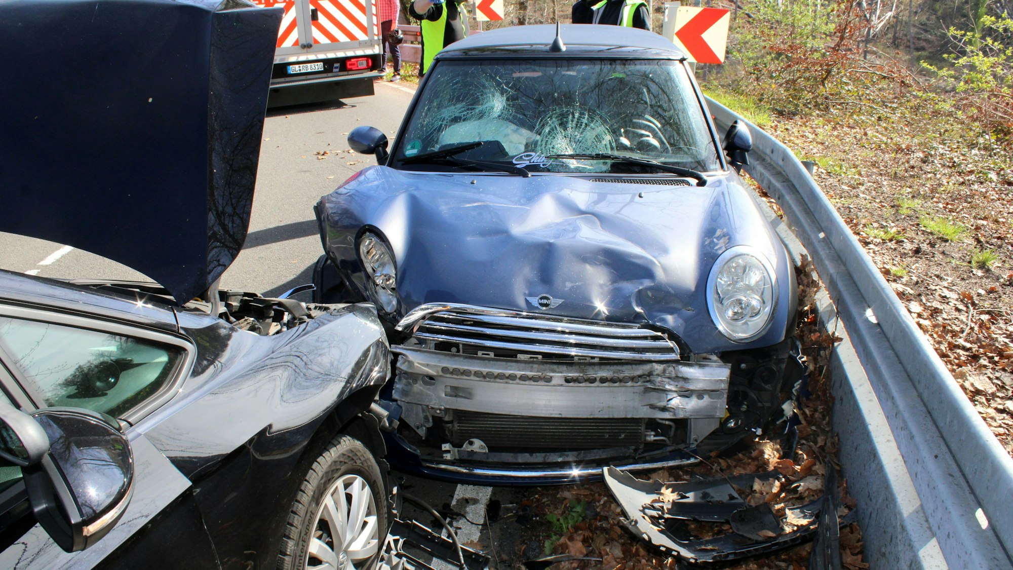 Zwei frontal zusammengestoßene Autos stehen an einer Leitplanke. Beide Fahrzeuge sind schwer beschädigt. Im Hintergrund stehen Einsatzkräfte von Polizei und Rettungsdienst.