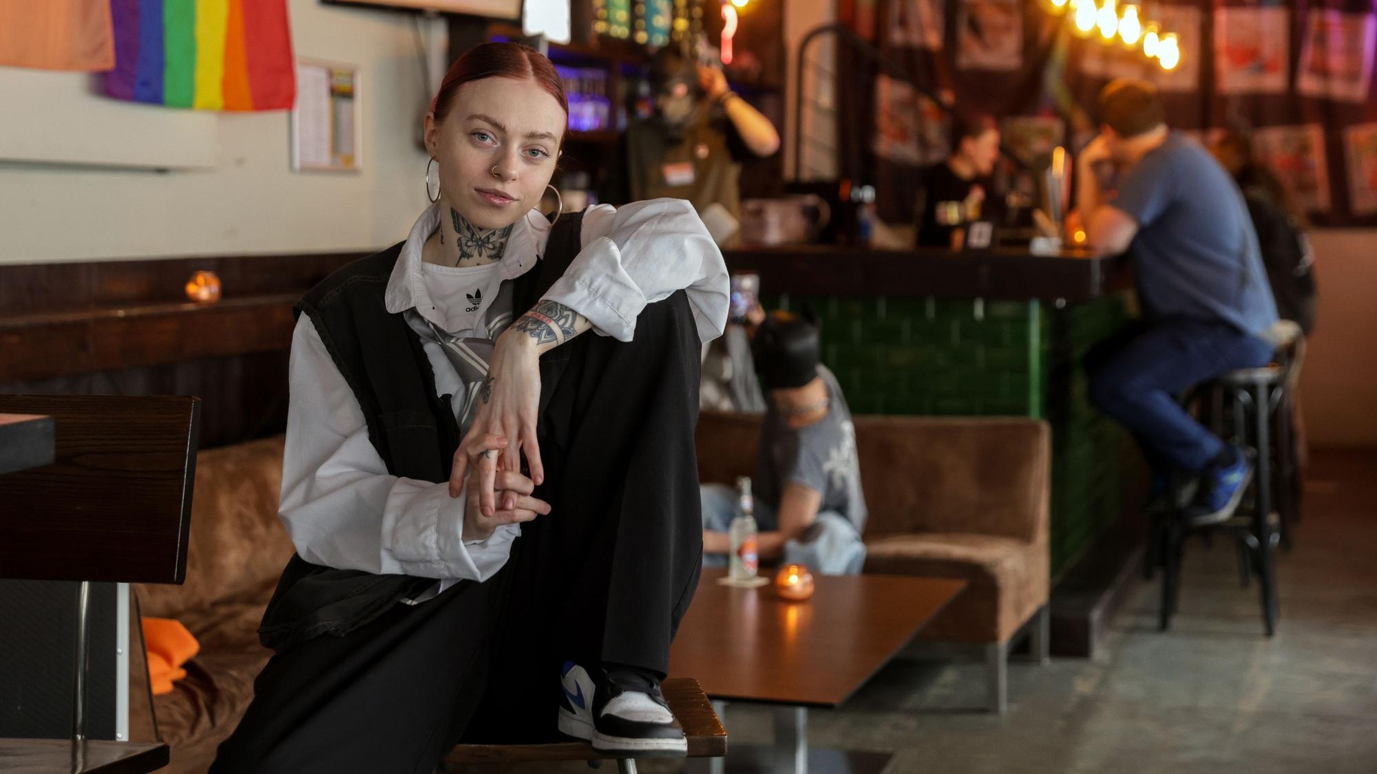 Influencerin Carina Hämmerle sitzt lässig im anyway, hinter ihr sind Leute an einer Bar, Lichter und Pride-Flaggen zu sehen. Hämmerle lächelt sanft in die Kamera.