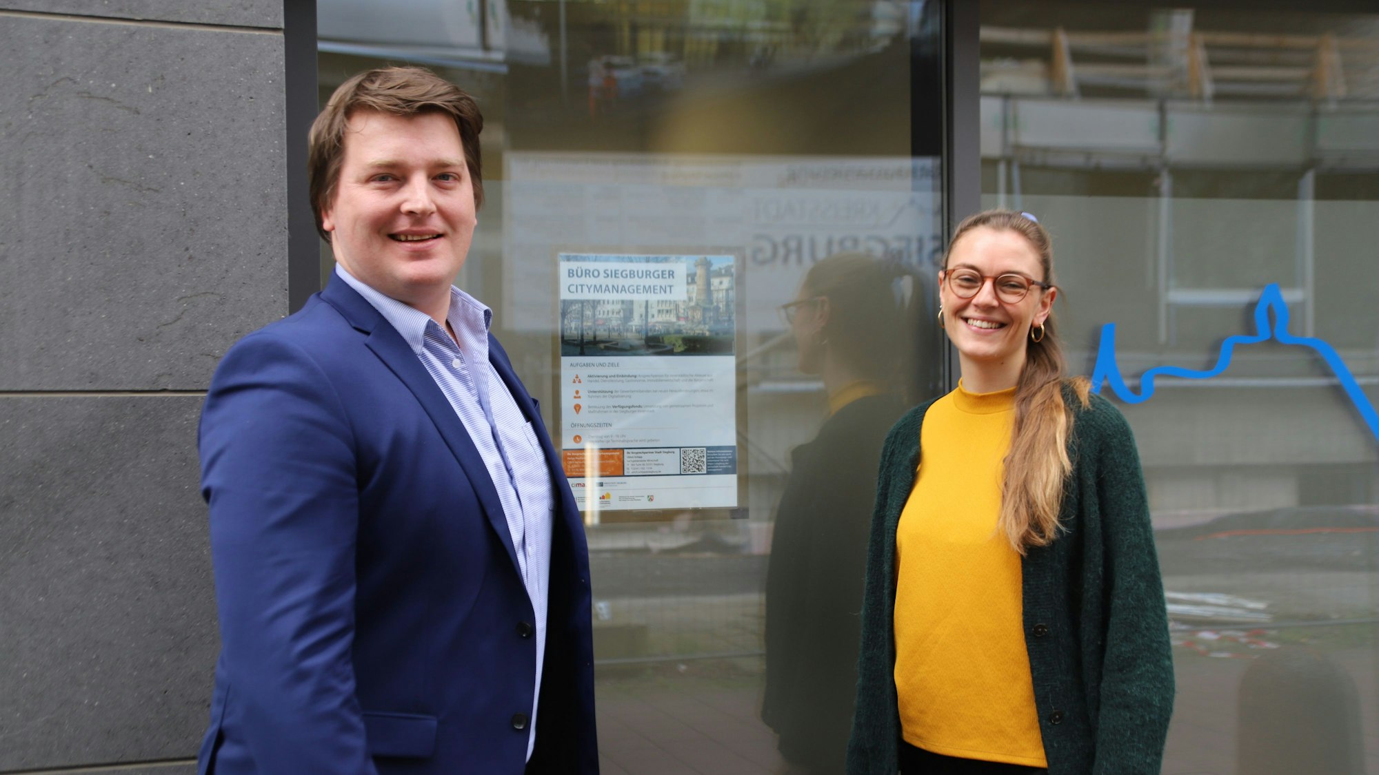 Helge Harnack (l.) und Amelie Strigl stehen vor der Tür zu ihrem Büro und lächeln in die Kamera.