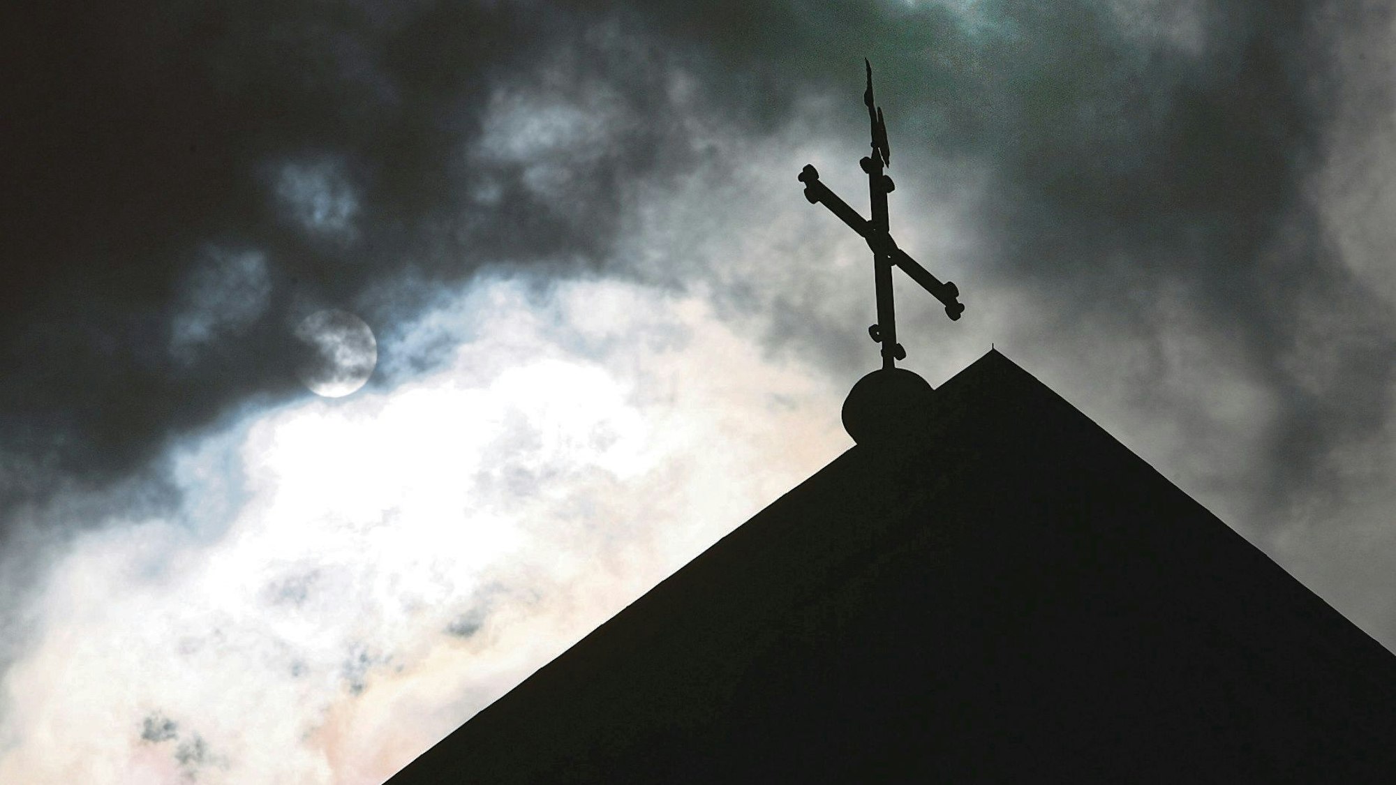 Im Gegenlicht und vor wolkenverhangenem Himmel ist die Kirchturmspitze des Doms mit Kreuz zu sehen.