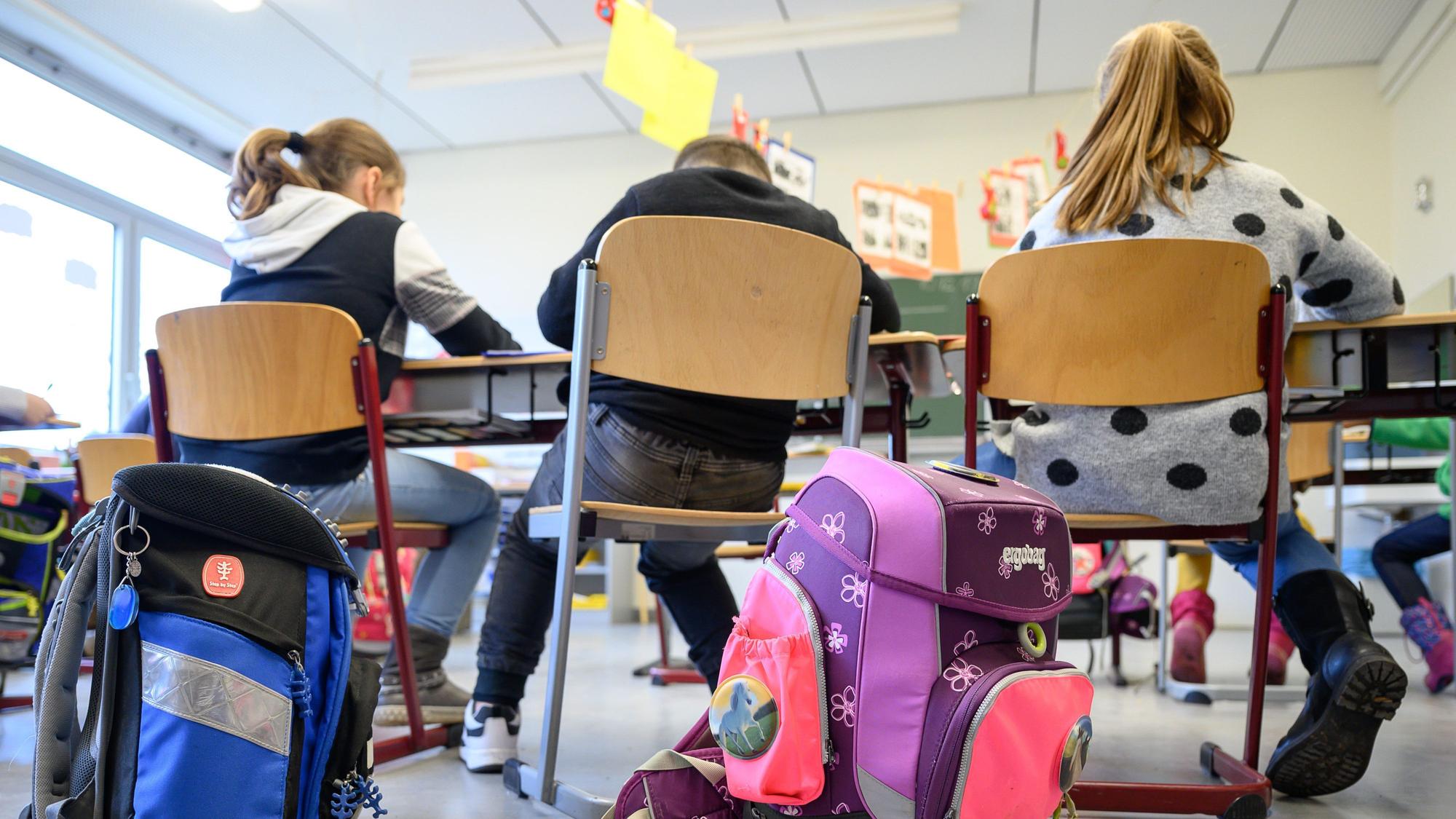 In einer Schulklasse sitzen Grundschüler auf ihren Stühlen. Im Vordergrund sind Ranzen zu sehen.