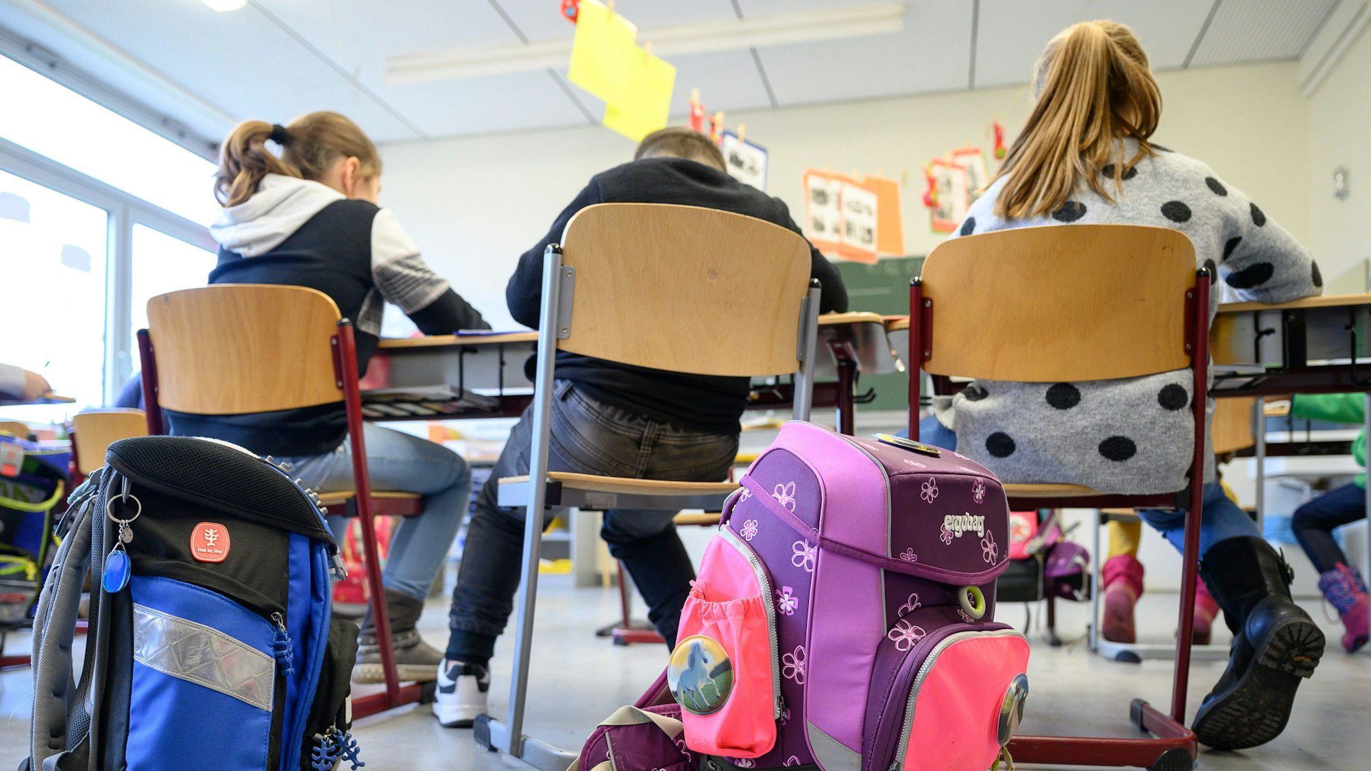 In einer Schulklasse sitzen Grundschüler auf ihren Stühlen. Im Vordergrund sind Ranzen zu sehen.