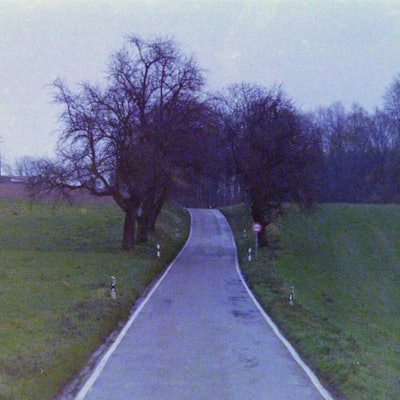Eine schmale asphaltierte Straße ohne Mittelstreifen schlängelt sich durch eine hügelige Landschaft, die von Ackerflächen und kleinen Waldstücken geprägt ist.