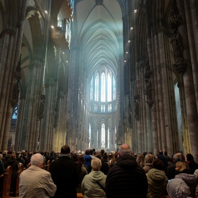 Blick in den Kölner Dom während einer Messe. Die Menschenmenge ist von hinten zu sehen, der Blick geht nach oben zu den gotischen Fenstern im Chor, die von Tageslicht erhellt sind.