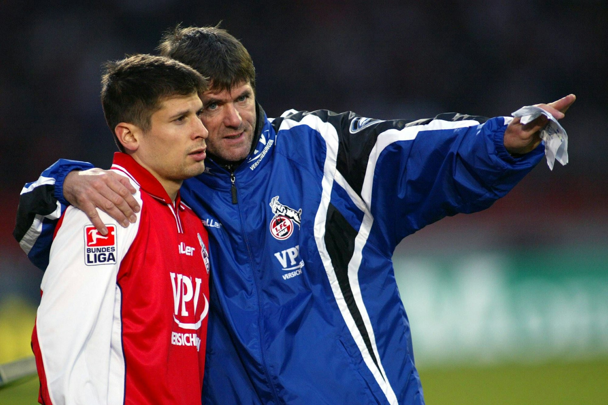 Ein Fußballspieler in rot-weißem Oberteil wird vom Trainer instruiert. Der Trainer in blauer Jacke hat den Arm um den Spieler gelegt.