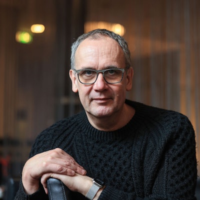 Kutscher sitzt in einem Café in Köln. Er trägt einen schwarzen Wollpulli und stützt sich mit den Händen auf eine Sitzlehne.