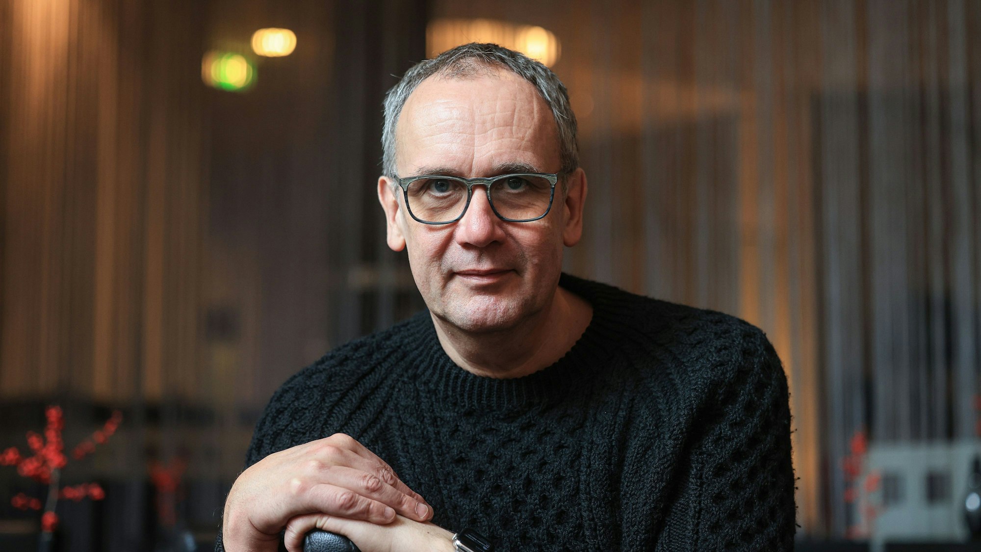 Kutscher sitzt in einem Café in Köln. Er trägt einen schwarzen Wollpulli und stützt sich mit den Händen auf eine Sitzlehne.