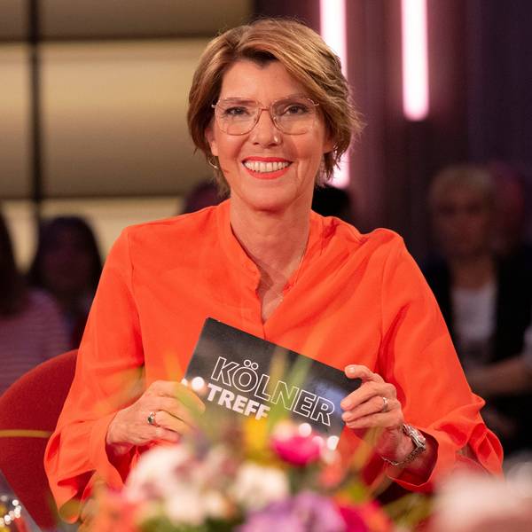 Moderatorin Bettina Böttinger, in orangefarbenem Kleid, hält ein Schild mit der Aufschrift „Kölner Treff“ in dei Kamera.