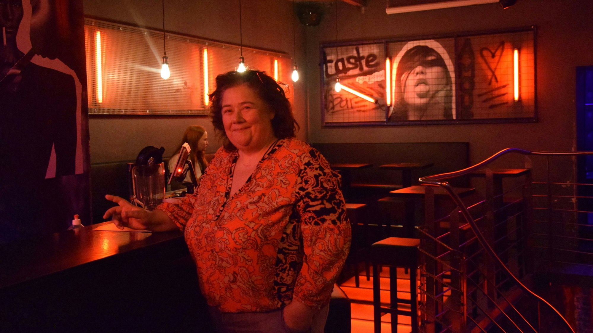 Ina Wolf steht an der Theke der BOIze.bar und lächelt in die Kamera. Hinter ihr sind Graffiti-Bilder, rote Lichter und Bartische mit Hockern.