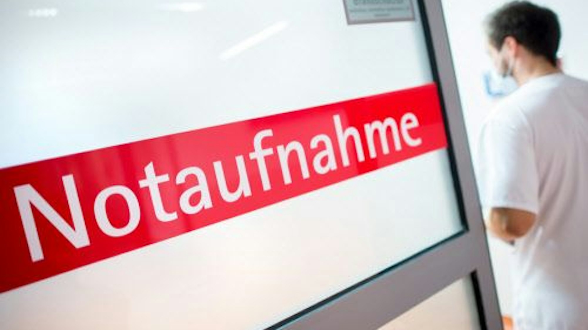 Ein Krankenpfleger steht in einem Krankenhaus neben einer Glastür mit der Aufschrift „Notaufnahme“