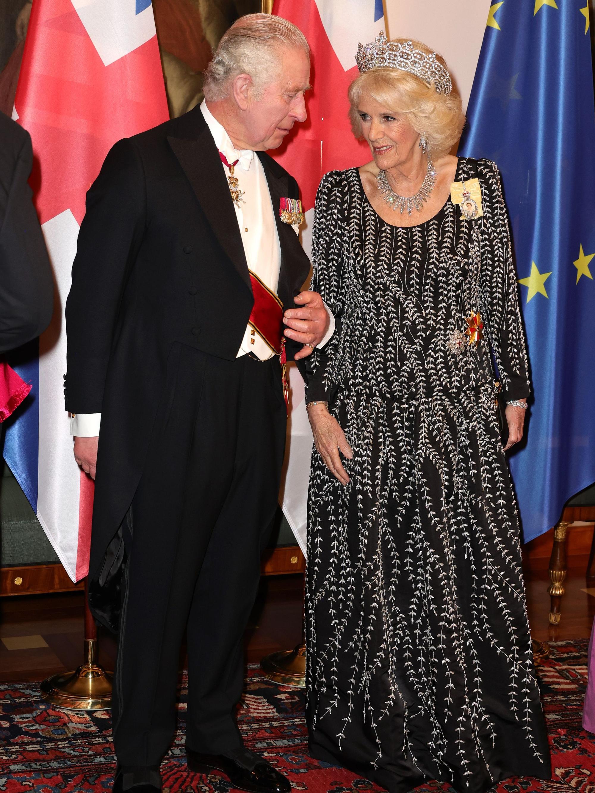 König Charles III. und Königin Camilla beim Staatsbesuch. Sie stehen vor Fahnen.