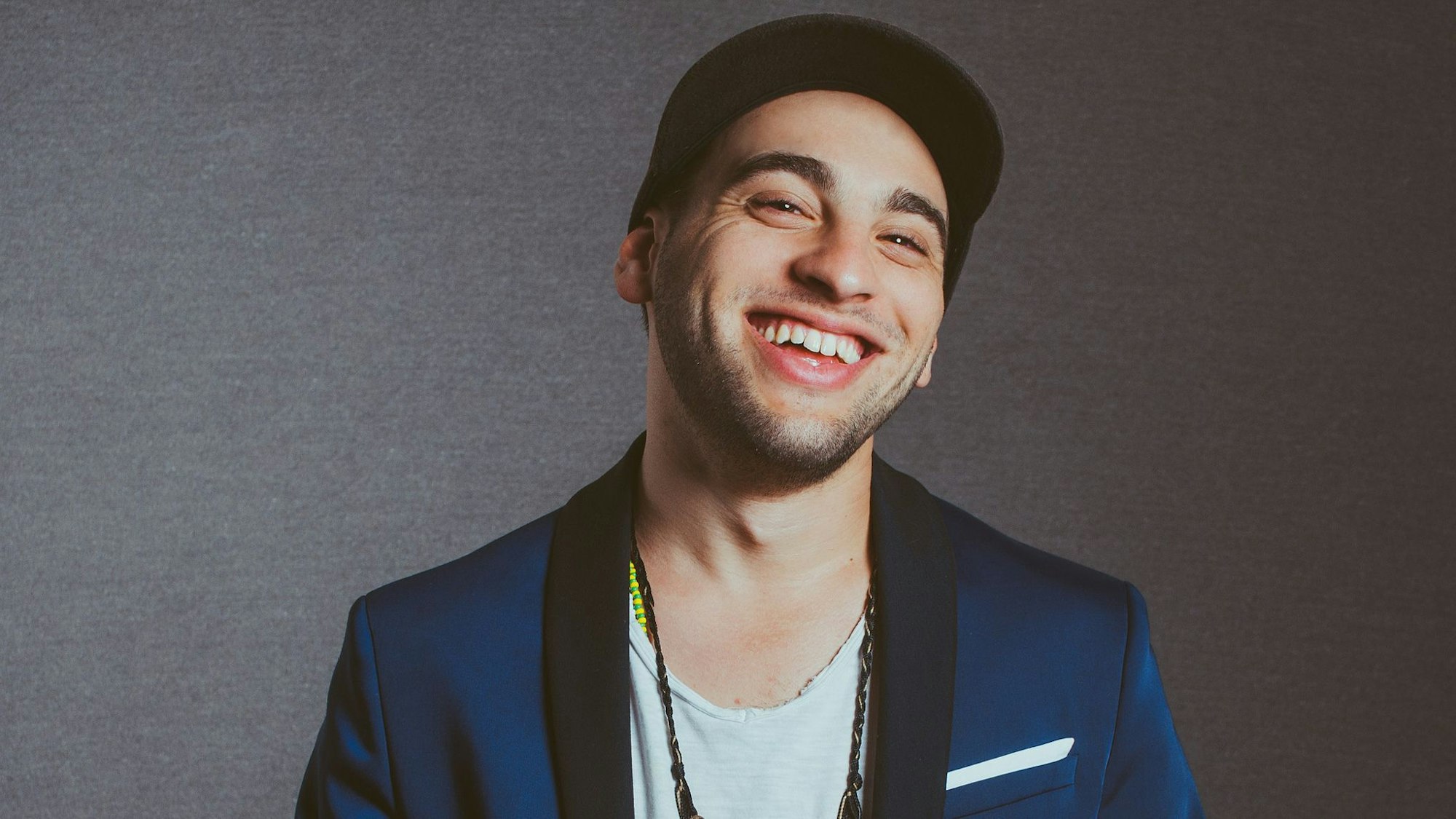 Samora Pinderhuges trägt eine Mütze, ein blaues Sacko und darunter ein weißes T-Shirt.