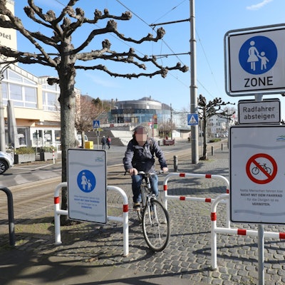 In Königswinter fährt ein Radfahrer trotz versetzter Sperren und Fußgängerzonen-Ausschilderung über die Rheinpromenade.