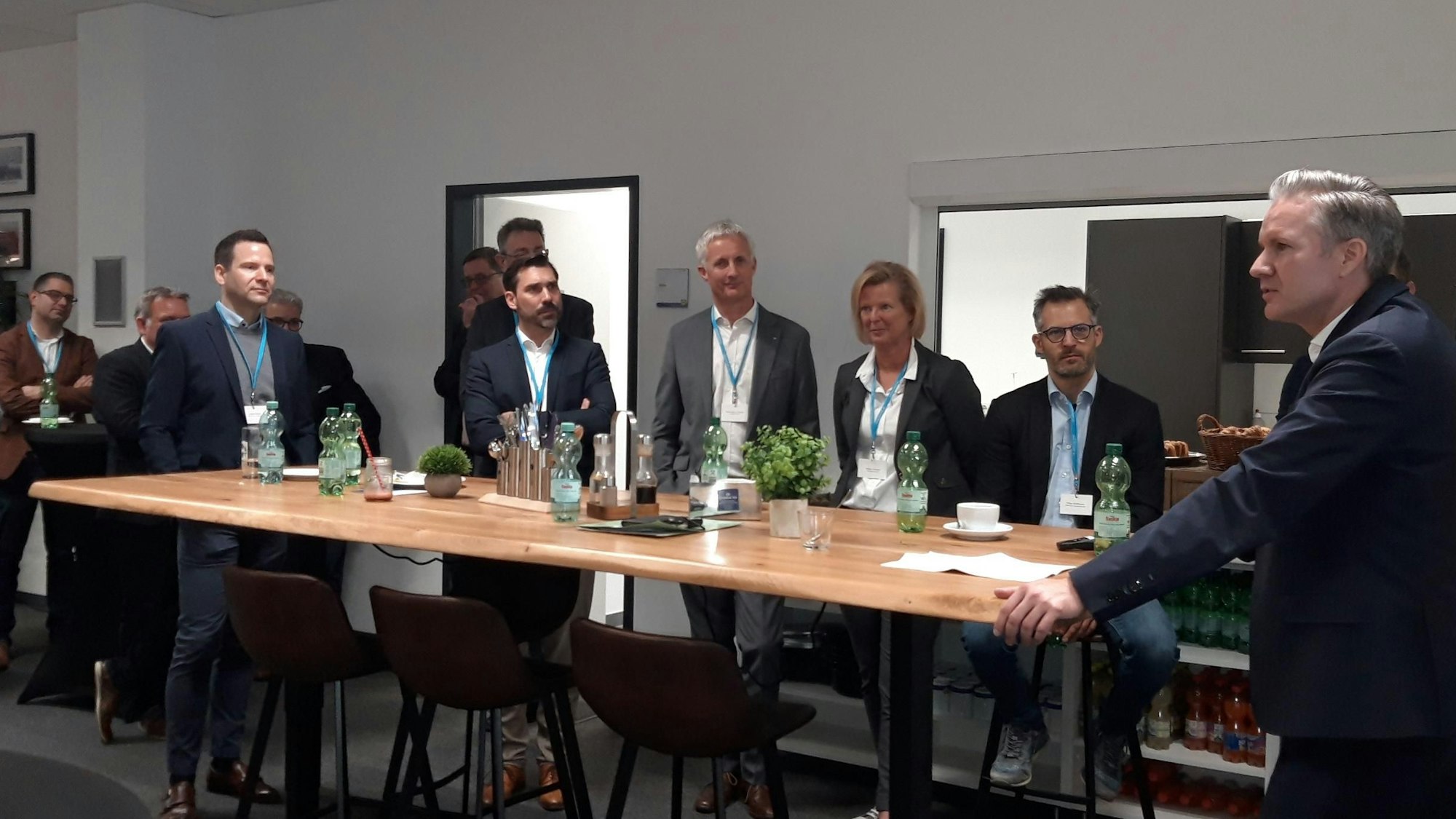 Am Kopfende eines langen Tisches steht mit aufgestützen Händen ein Mann, an der Längsseite des Tisches stehen mehrere Männer und Frauen und hören ihm zu.