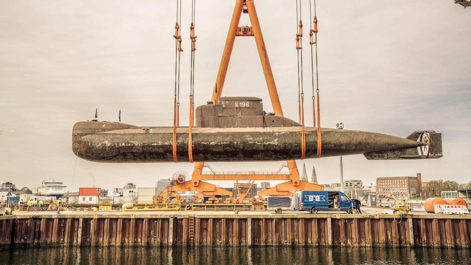 Ein großes U-Boot hängt in Schlingen an einem großen Kran und hängt über einem Seedock.