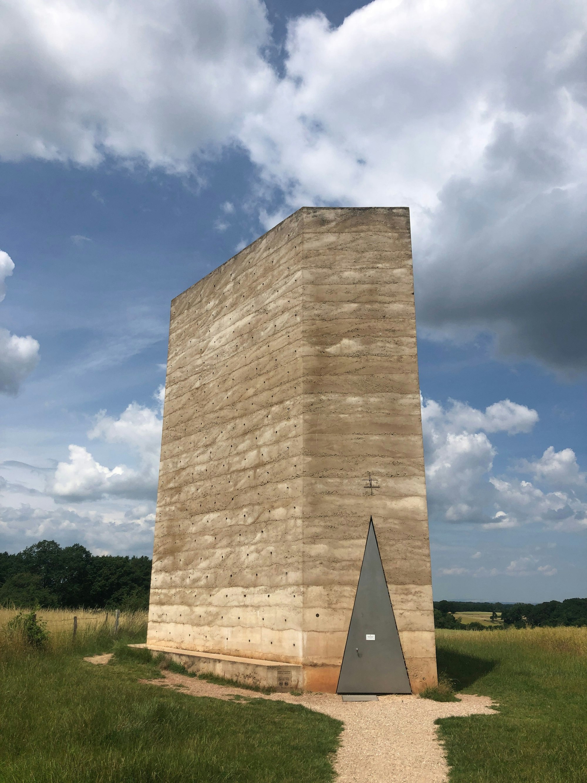 Die Bruder Klaus Kapelle von Peter Zumthor steht mit Dreieckstür vor einem blauen Himmel.