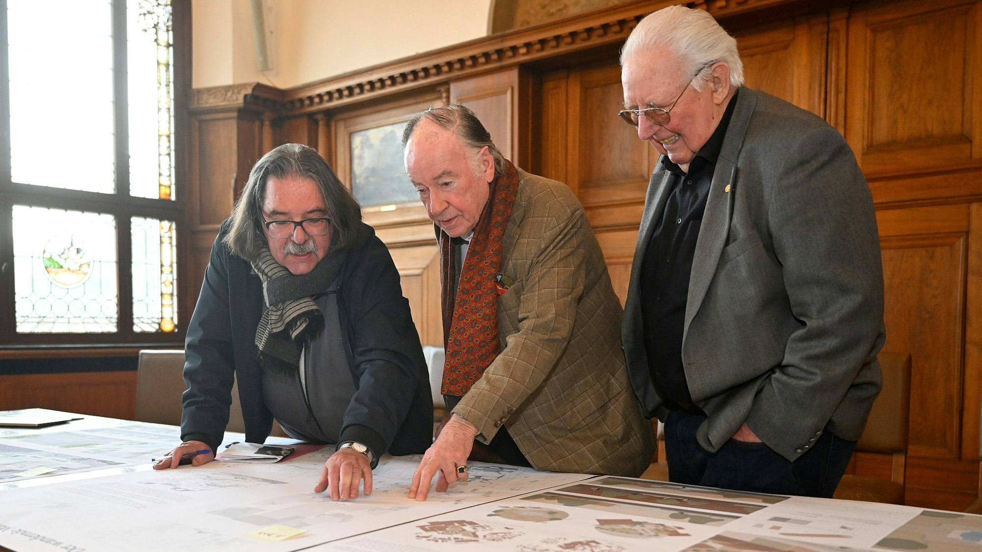 Drei ältere Herren zeigen auf einen ausgebreiteten Entwurf, der auf einem Tisch liegt.