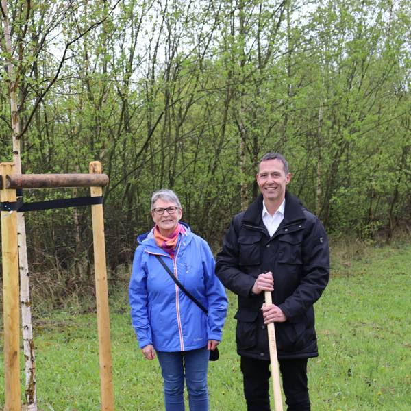 Die drei Personen stehen auf einer Wiese um den jungen Baum herum.