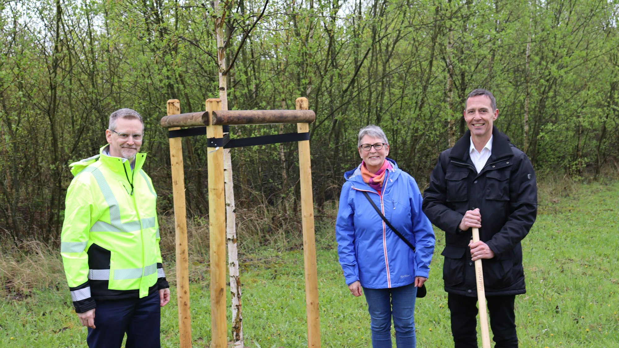 Die drei Personen stehen auf einer Wiese um den jungen Baum herum.