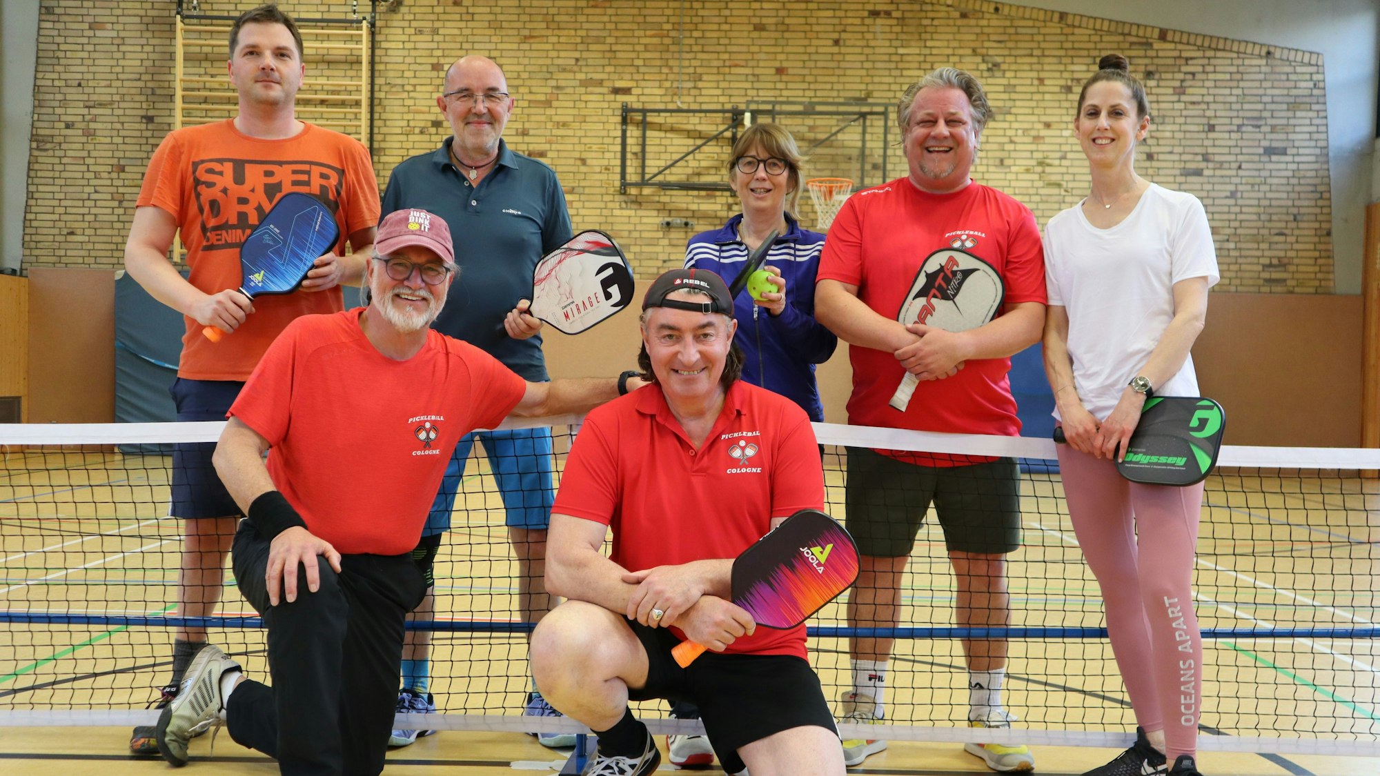 Fünf Männer und zwei Frauen unterschiedlichen Alters in Sportkleidung in einer Turnhalle vor einem Netz mit Schlägern in der Hand