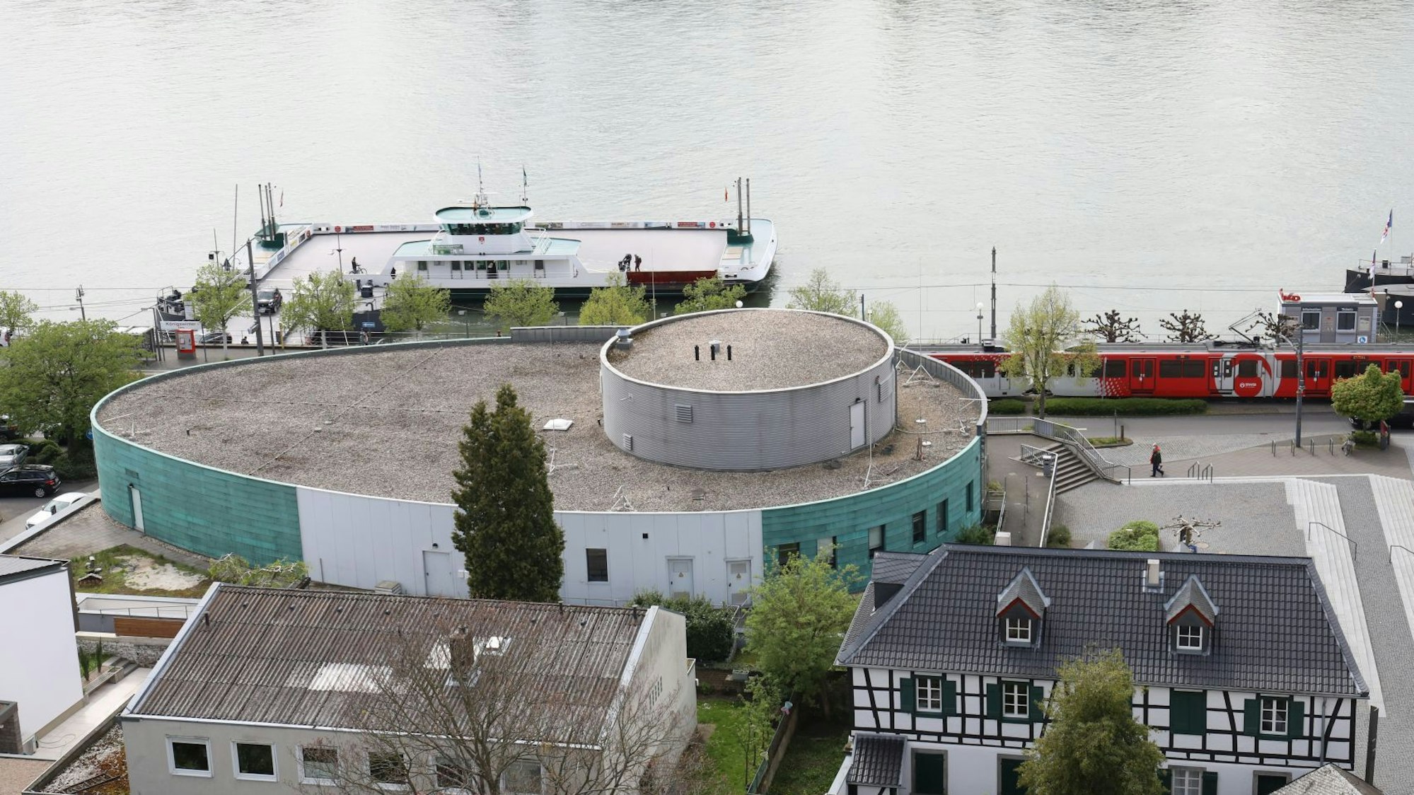 Hinter dem ovalen Gebäude ist die Rheinfähre Königswinter zu sehen, rechts steht eine Stadtbahn der Linie 66.