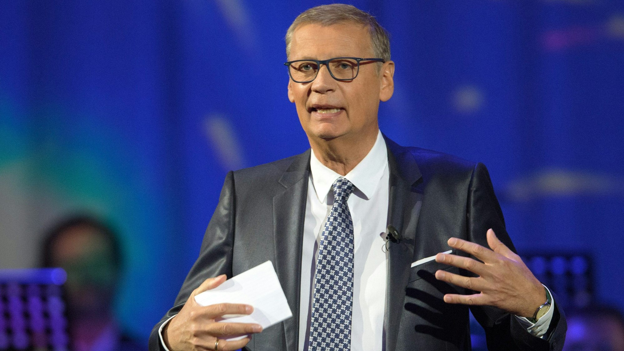 „Wer wird Millionär?“-Moderator Günther Jauch sitzt auf einer Bühne. In der Hand hält der RTL-Moderator eine Karte.