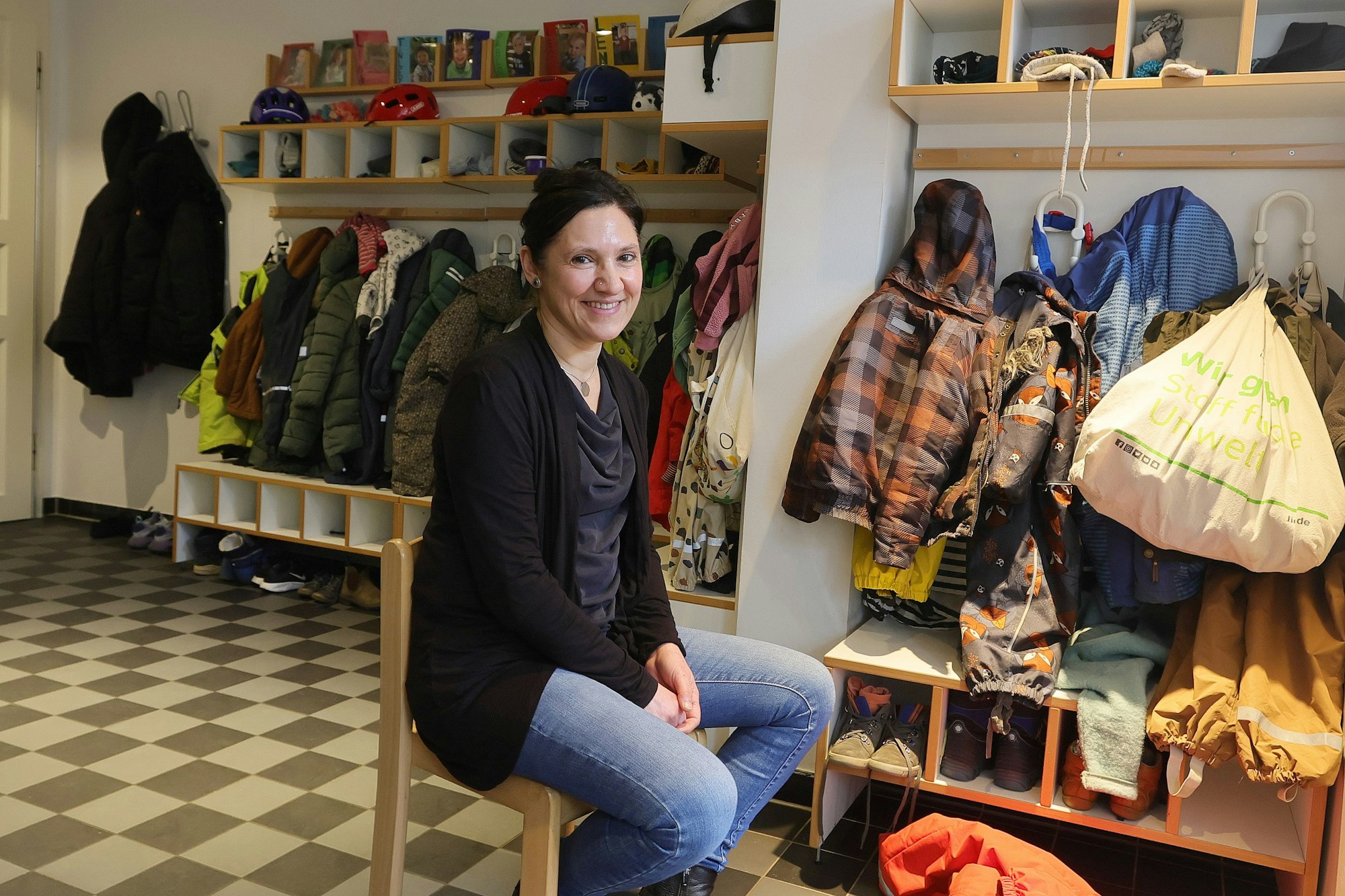Die Kita-Leiterin Tatjana Murgia sitzt auf einem Stuhl vor der Kita-Garderobe und vielen Jacken.