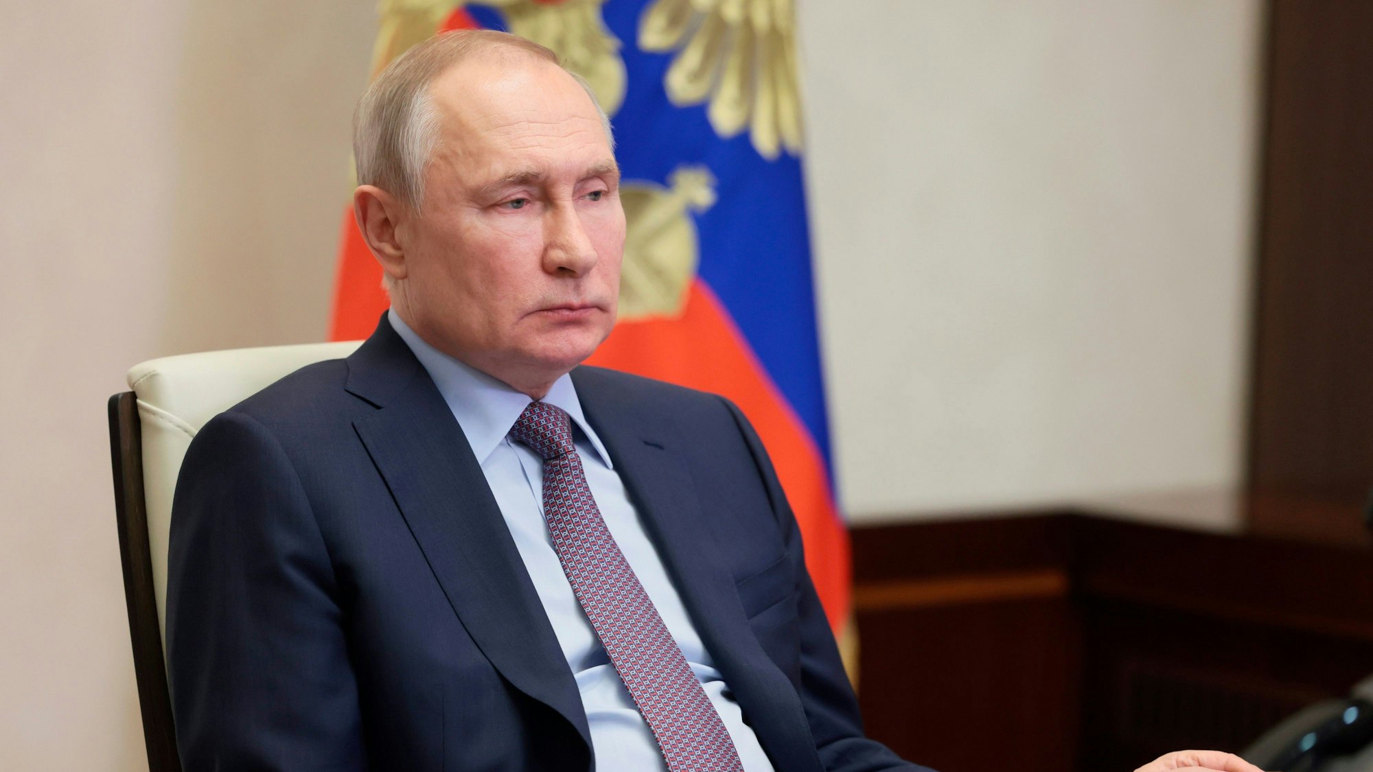 Der russische Präsident Putin sitzt auf einem Stuhl. Hinter ihm steht eine Flagge.