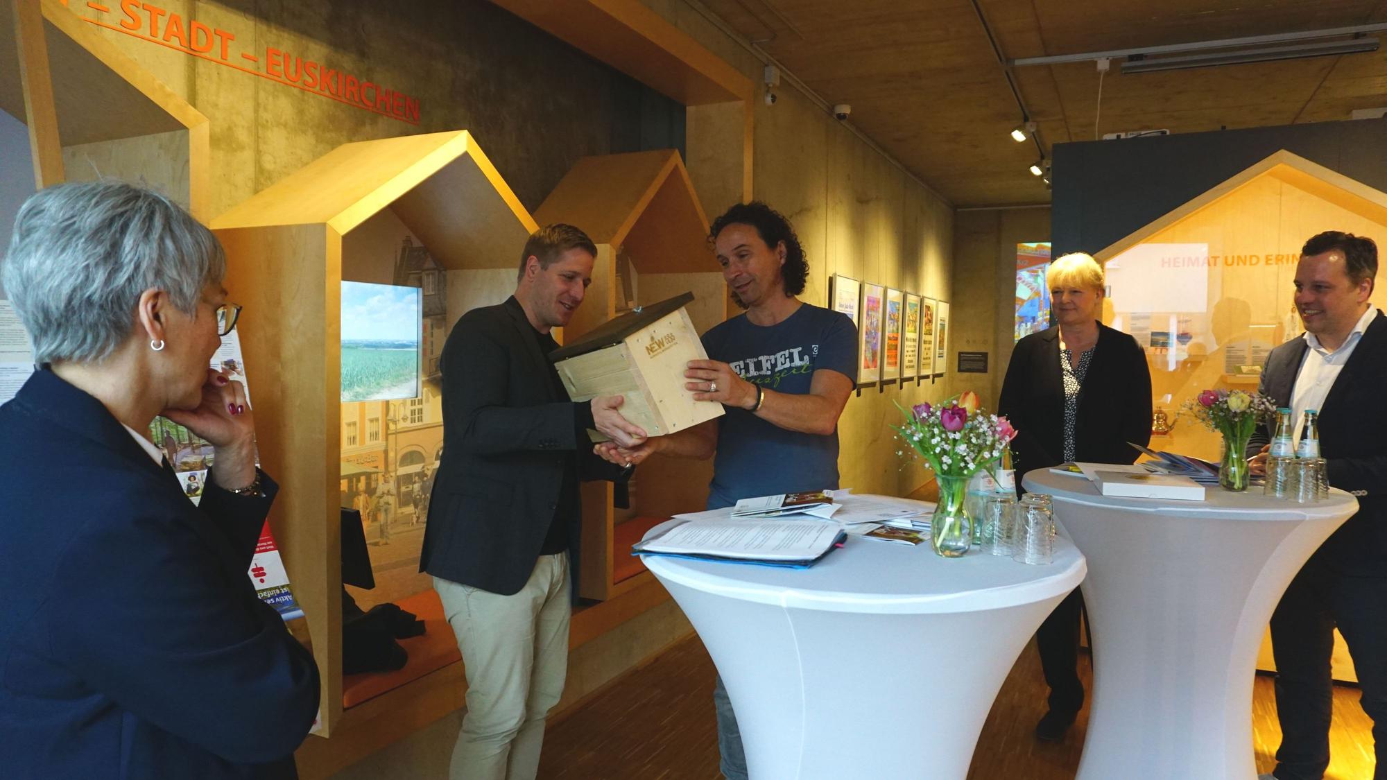 Landrat Markus Ramers überreicht Stefan Brings, dem Paten der Aktion „Zu Gast in der eigenen Heimat“ ein „Hummelhaus“. Um sie herum drei weitere Personen. Sie befinden sich drinnen.