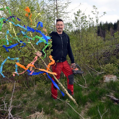 Ein junger Mann steht im Wald und hält einen mit bunten Bändern geschmückten Maibaum in der Hand. In der anderen Hand hält er eine Motorsäge.