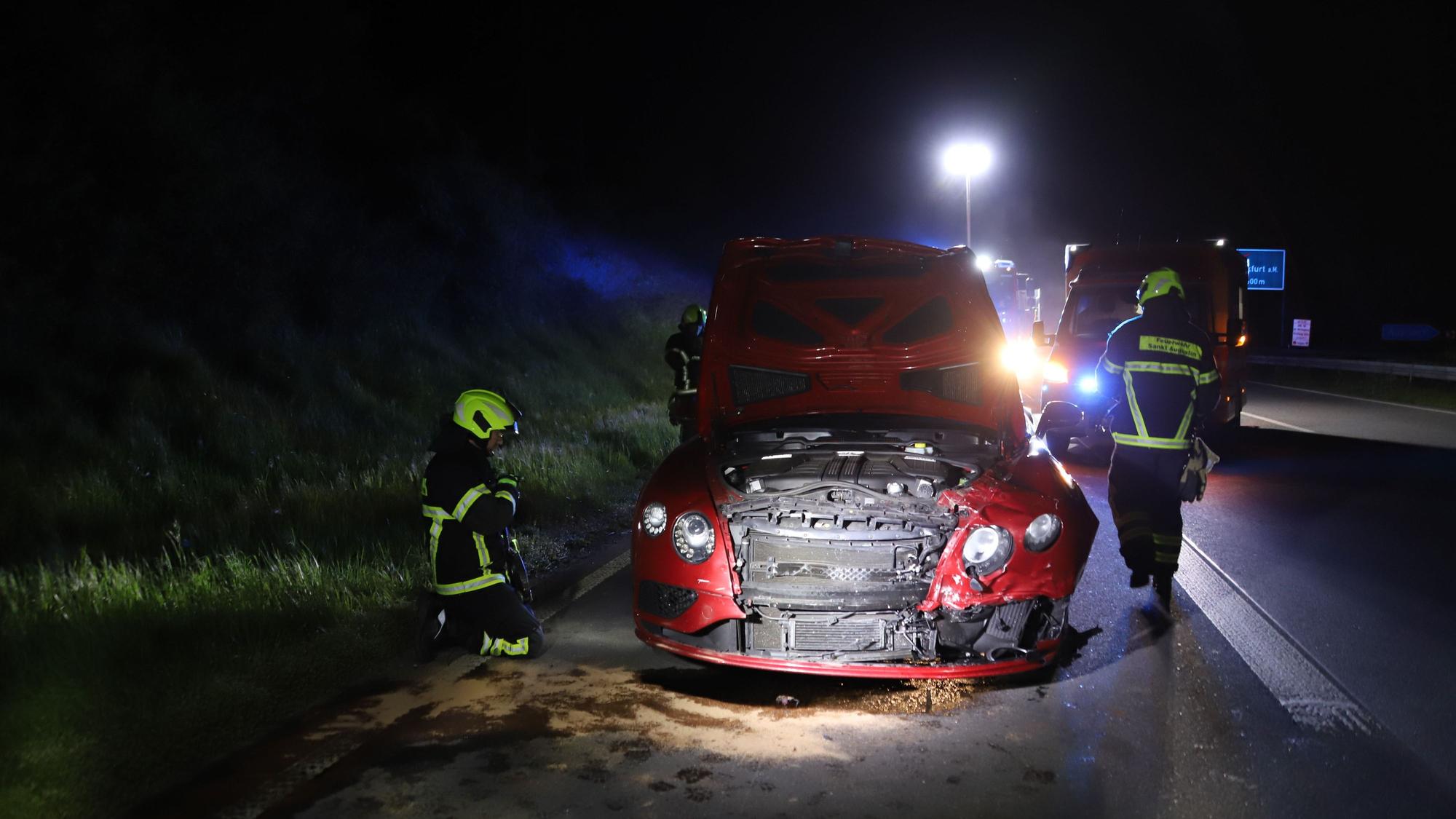 Feuerwehrkräfte begutachten nachts ein rotes Auto nach einem Unfall. Die Front ist von einer Kollision beschädigt.
