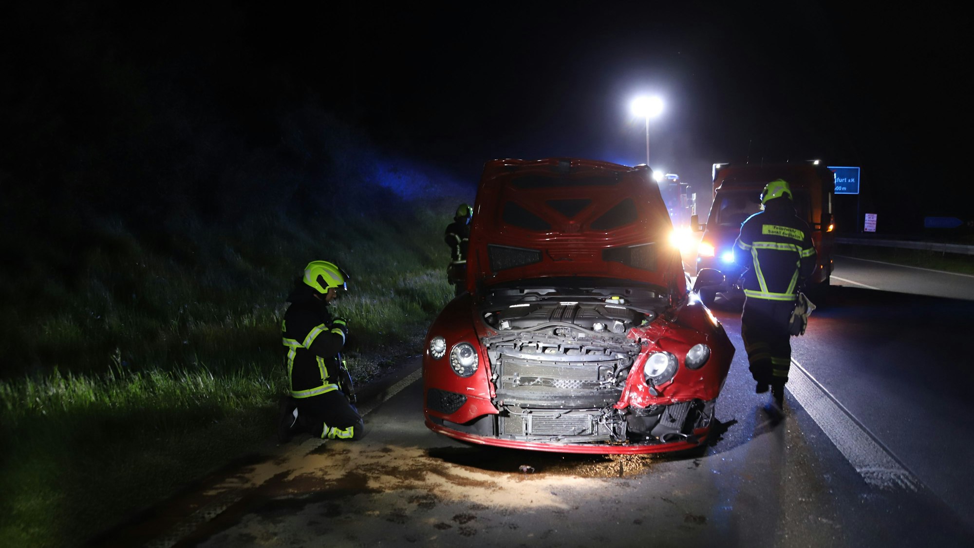 Feuerwehrkräfte begutachten nachts ein rotes Auto nach einem Unfall. Die Front ist von einer Kollision beschädigt.