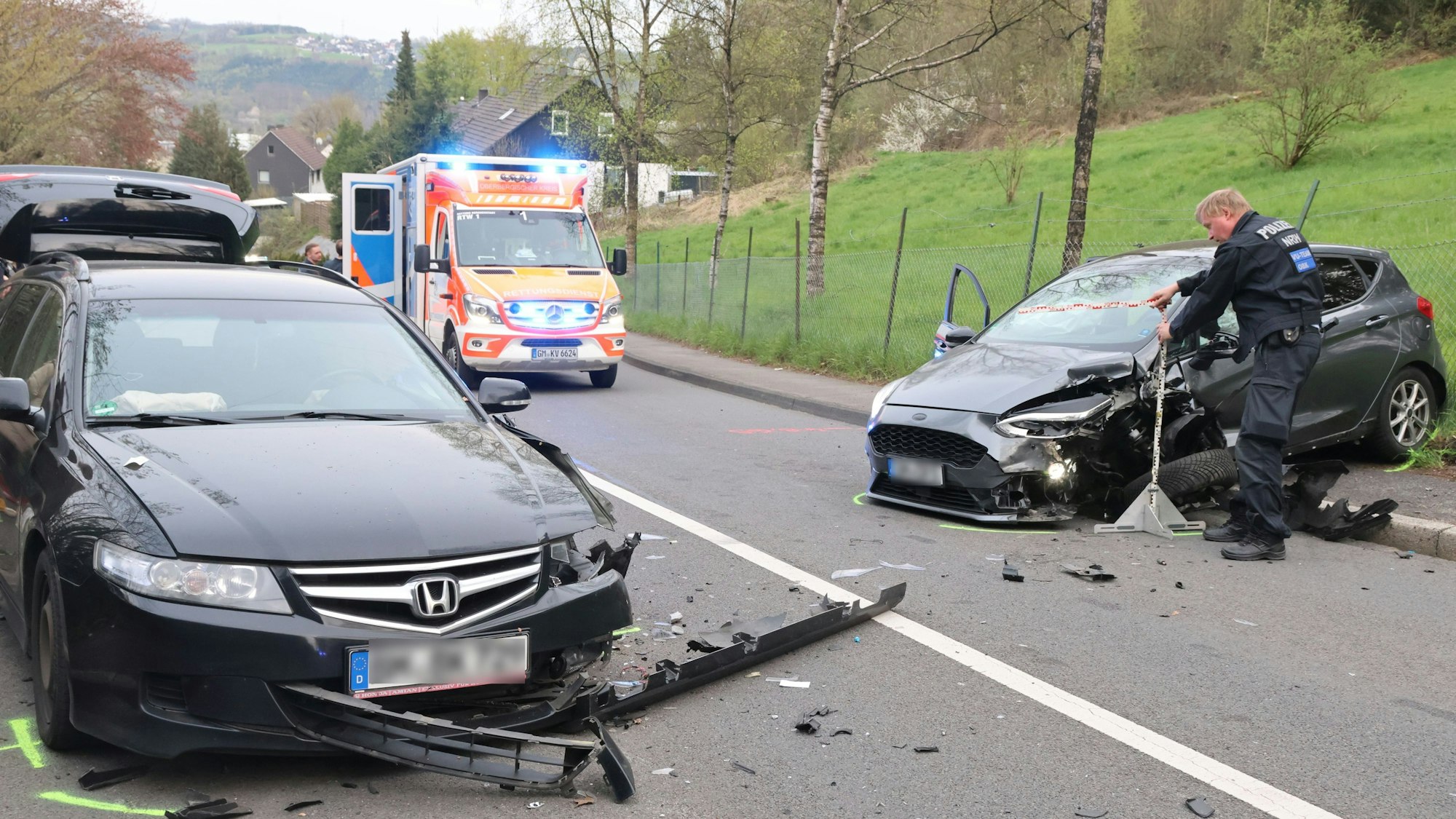 Zwei nach einer Kollision beschädigte Fahrzeuge stehen auf einer Straße. Ein Polizist fegt die Scherben von der Straße. Im Hintergrund steht ein Krankenwagen.