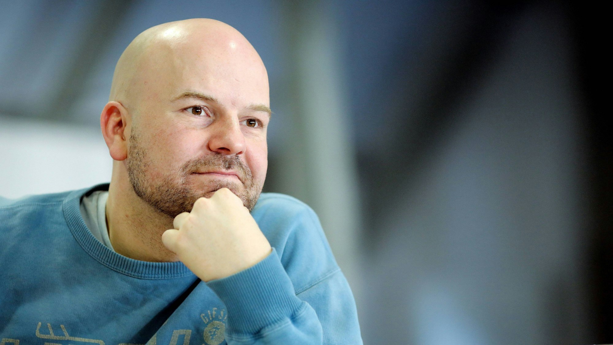 Manuel Moser stützt sich das Kinn mit der Hand. Er hat eine Glatze und einen 3-Tage-Bart. Er trägt einen blauen Pulli.