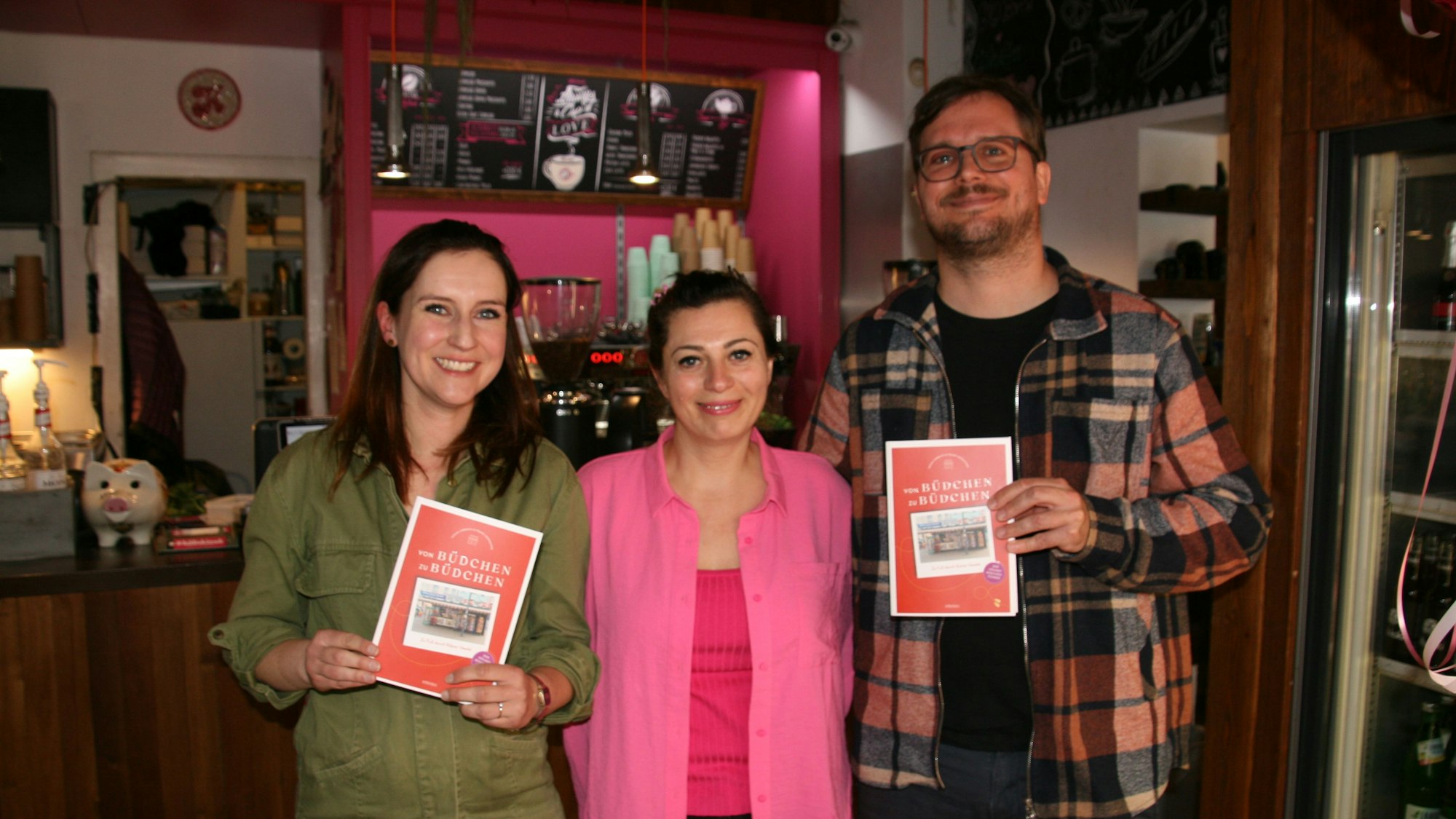 Tanja Limbach, Shirin Shaghaghi und Frank Münschke stehen in einem Büdchen. Die beiden Autoren haben den Kölner Büdchen-Führer in der Hand.