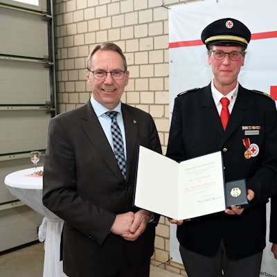 Sascha Habernickel steht in Uniform zwischen zwei Männern im Anzug und hält lächelnd seine Auszeichnung und Medaille in die Kamera.