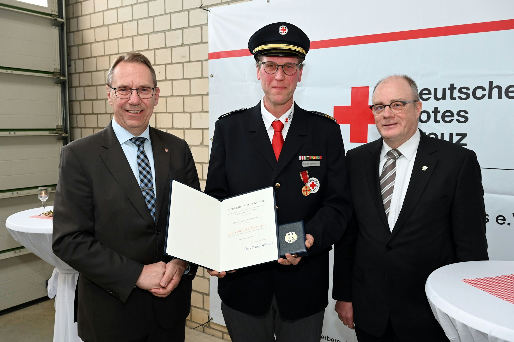 Sascha Habernickel steht in Uniform zwischen zwei Männern im Anzug und hält lächelnd seine Auszeichnung und Medaille in die Kamera.