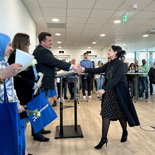 Ein Mann im dunklen Sakko schüttelt einer Frau mit schwarzen Haaren und ebenfalls dunkler Kleidung die Hand. Beide lächeln.