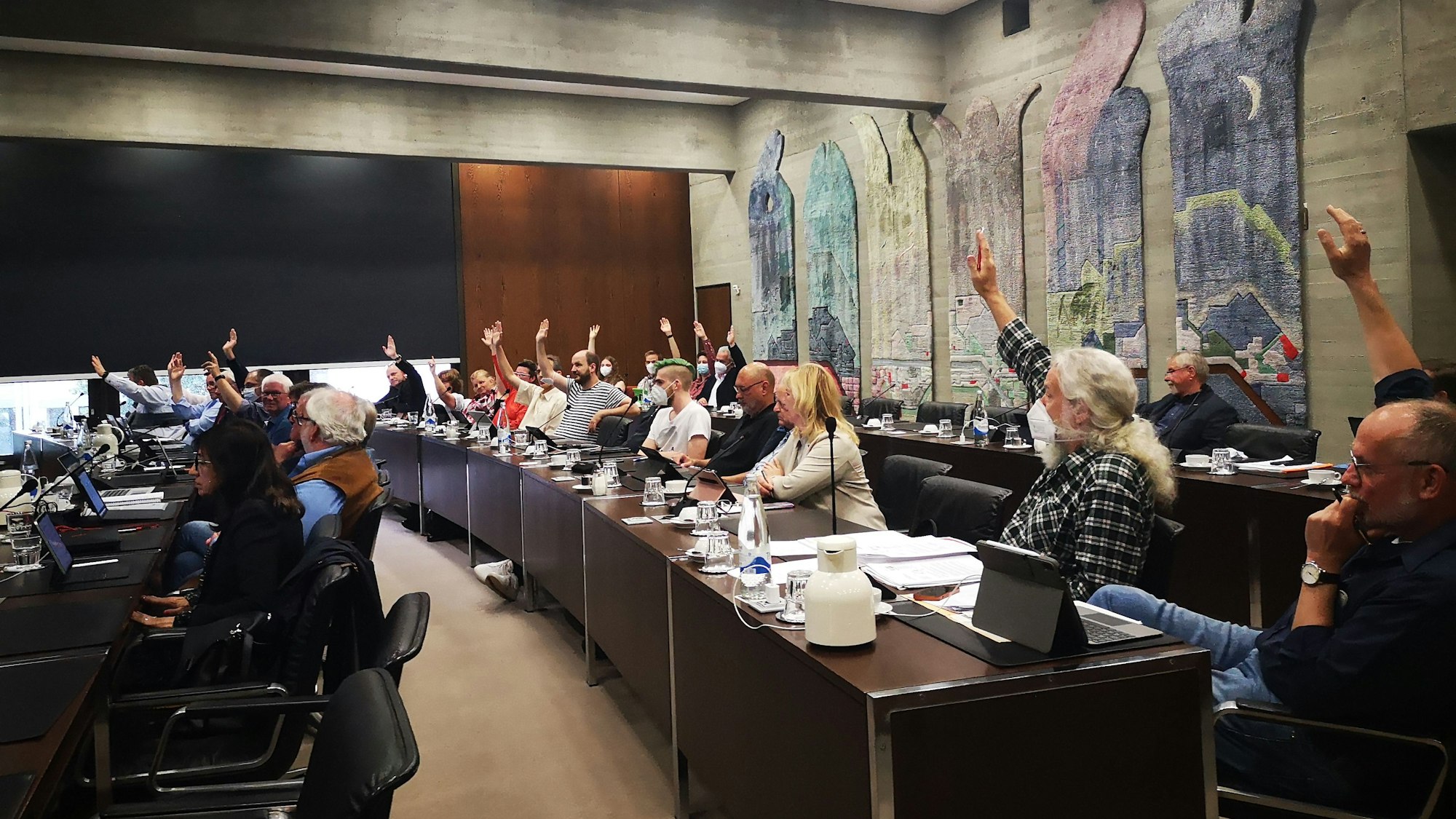 Die Ratsmitglieder in Wesseling stimmen im Ratssaal über Beschlüsse ab. Die Menschen sitzen an langen Tischen und heben teilweise ihrer Hand zur Abstimmung.