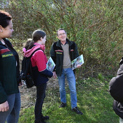 Ein Mann steht mit mehreren Schülerinnen im Wald. Er hat eine Informationsmappe in der Hand und spricht zu den jungen Frauen.
