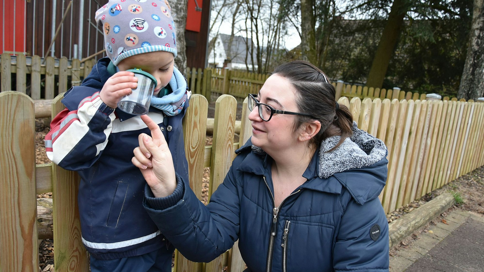 Ein kleines Kind schaut mit einem Auge in ein Glas. Eine Frau kniet neben dem Kind und zeigt auf den Inhalt des Glases.
